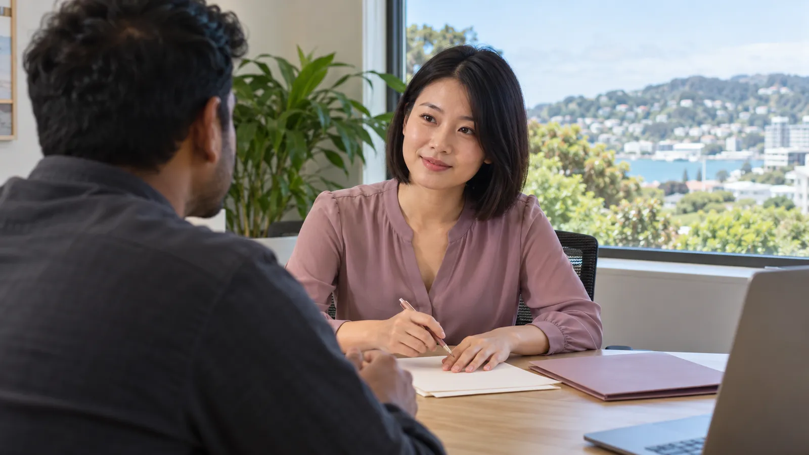 A woman in mauve discusses paperwork with a colleague in a sunlit office overlooking a harbour.