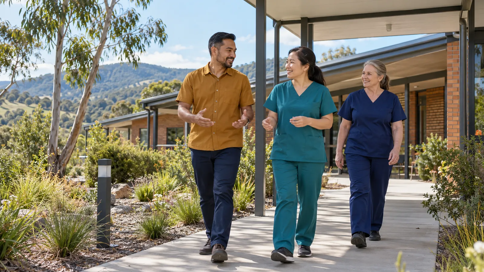 A man in an ochre shirt walks and chats with two healthcare colleagues outside a leafy regional clinic.