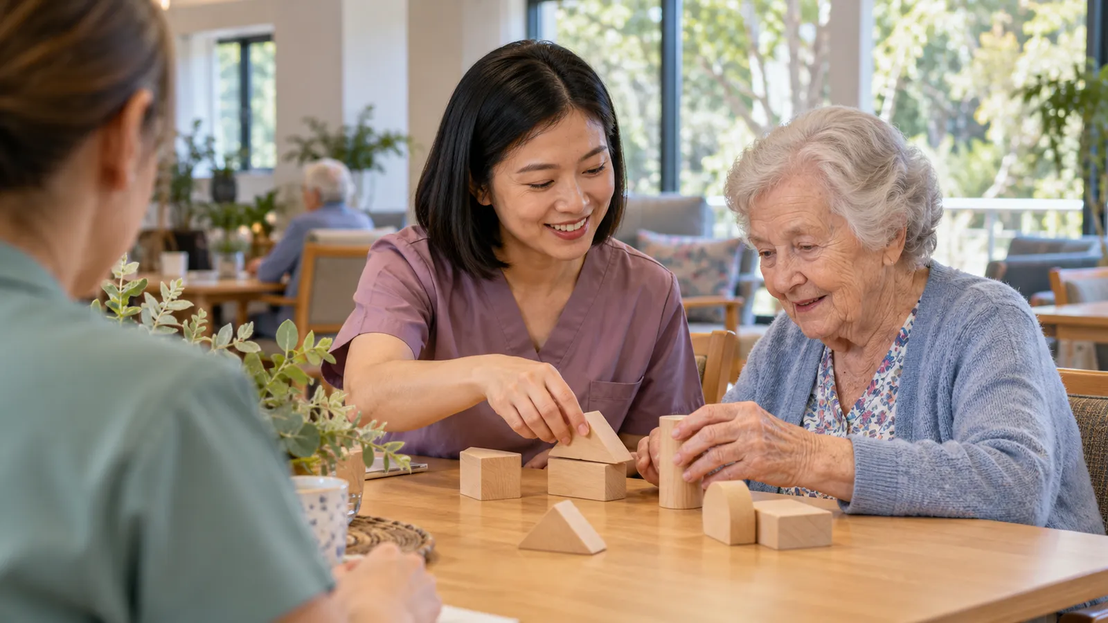 A care worker in mauve scrubs helps an elderly woman arrange wooden blocks as a colleague looks on.