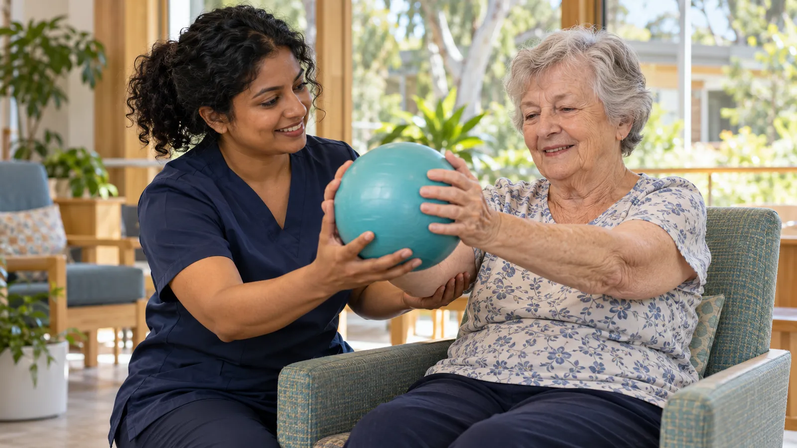 A care worker in navy helps an older woman exercise with a turquoise ball in a sunlit care room.