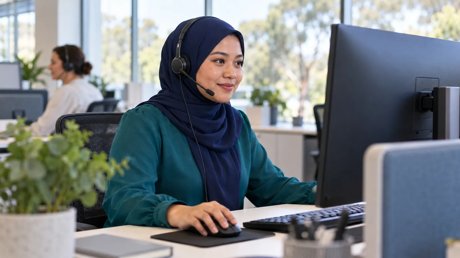 A woman in a navy hijab and teal blouse uses a headset at her desk in a bright customer care office.